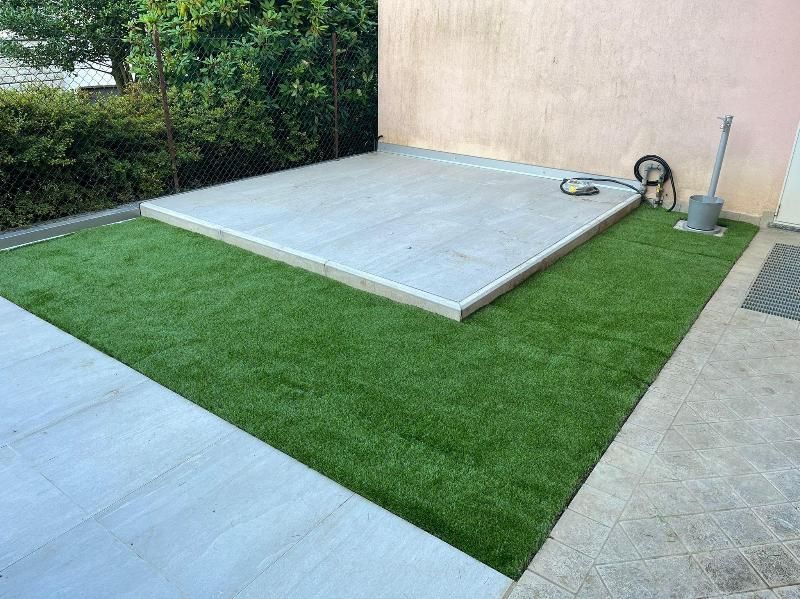 Piccolo patio in cemento e zona con erba sintetica accanto a un muro beige, con un tubo da giardino e una pala nelle vicinanze.