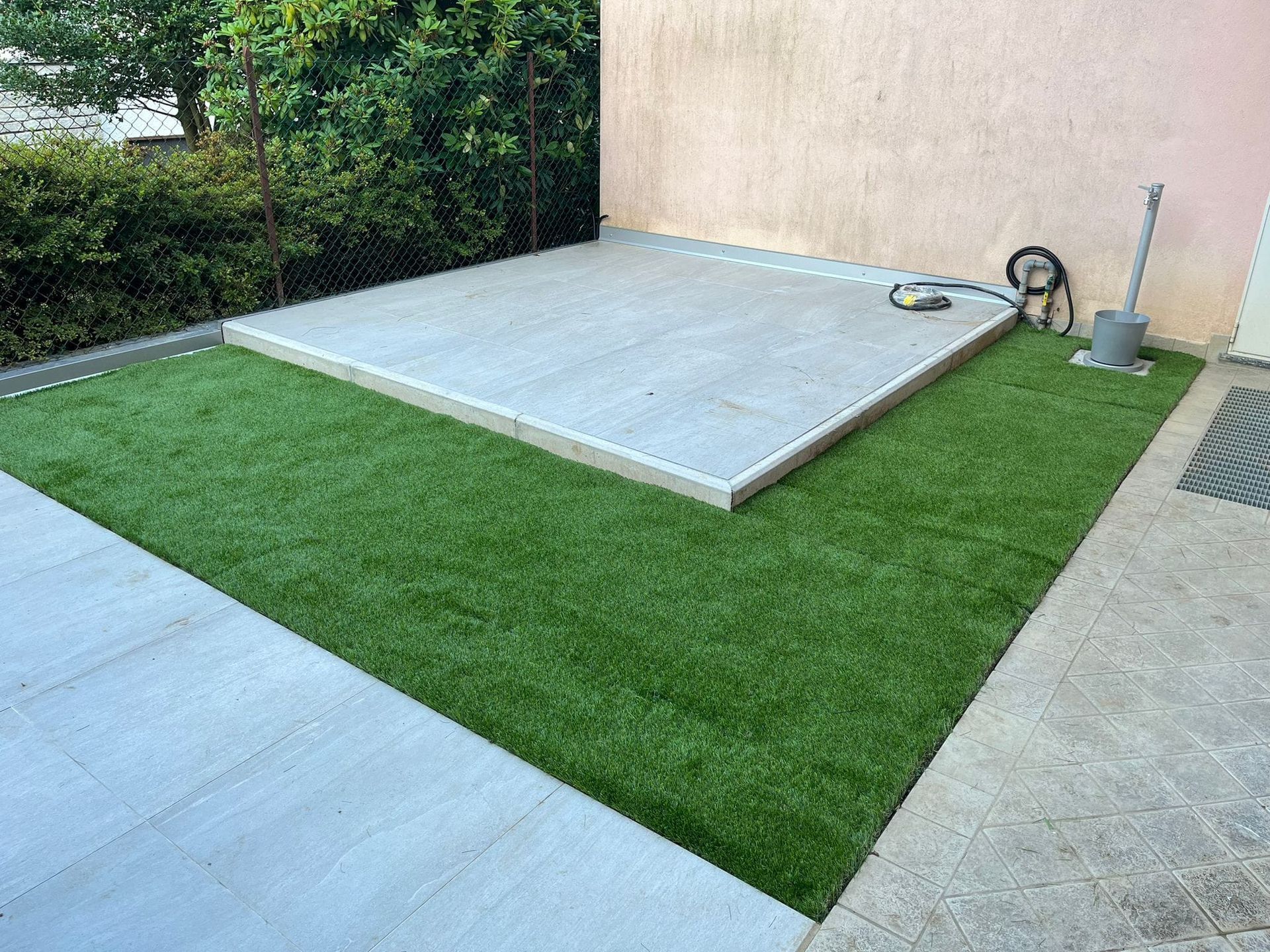 Piccolo patio sul retro della casa con una piattaforma rialzata in cemento accanto a un prato verde e un muro beige.