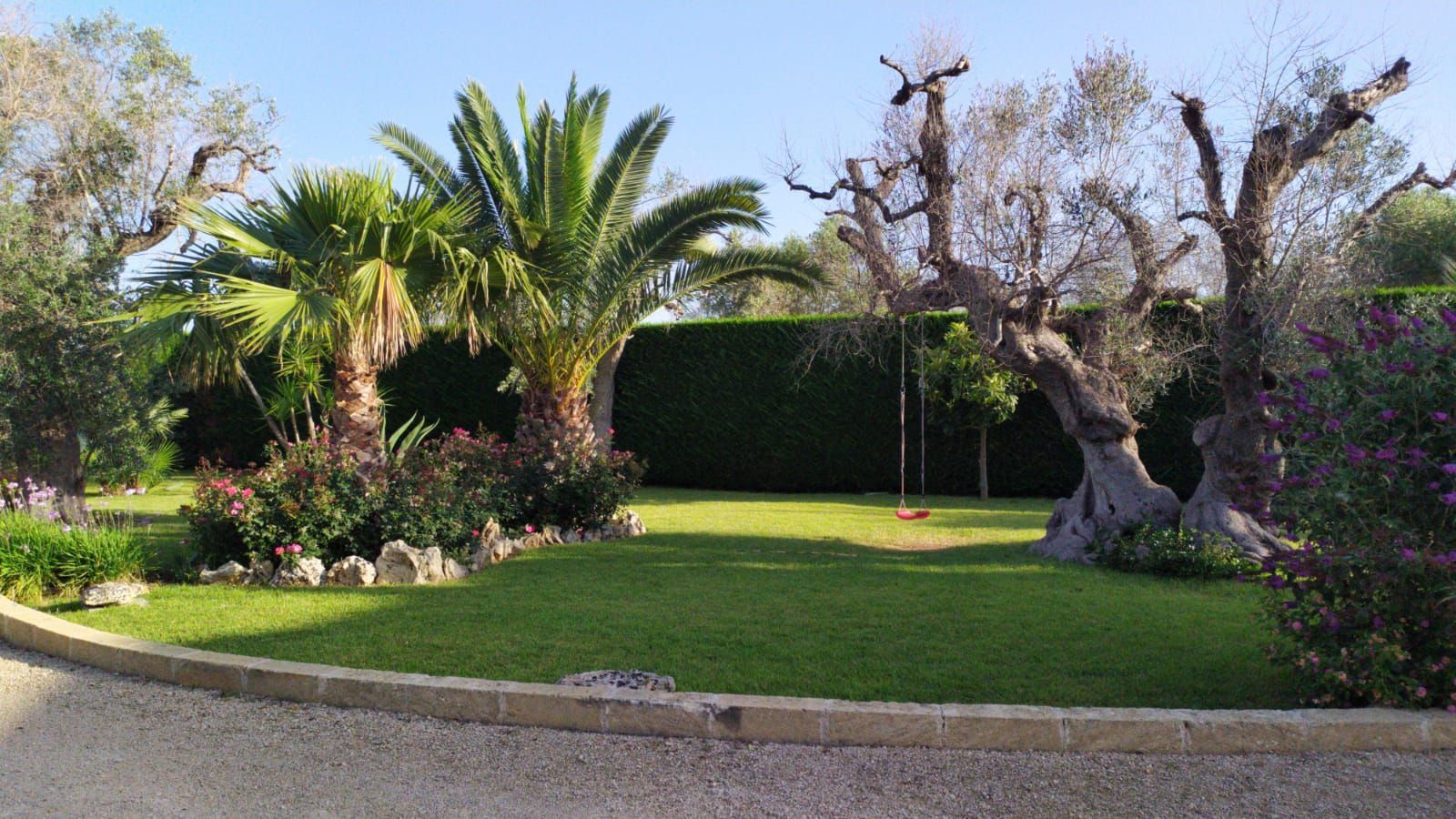 Un giardino lussureggiante con palme, un grande albero nodoso, un prato verde e arbusti fioriti in piena luce del sole.
