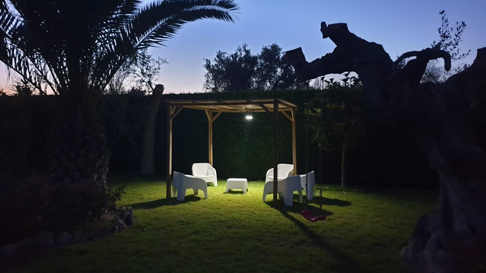 Gazebo in giardino illuminato al crepuscolo, con sedie bianche e un rigoglioso prato verde stagliato contro le palme.