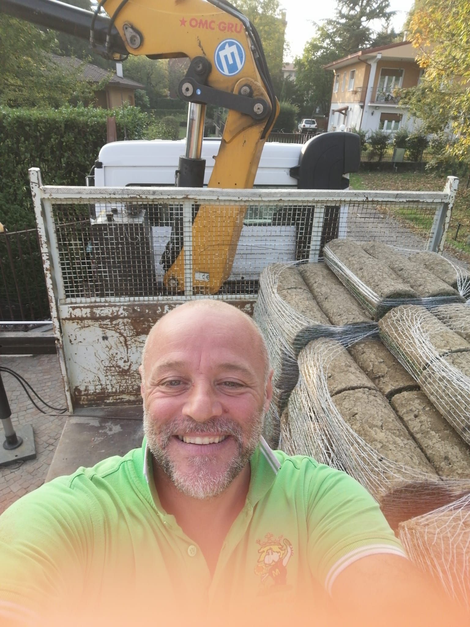 Un uomo sorridente con una maglietta verde si scatta un selfie in un cantiere edile, con un escavatore giallo alle sue spalle.