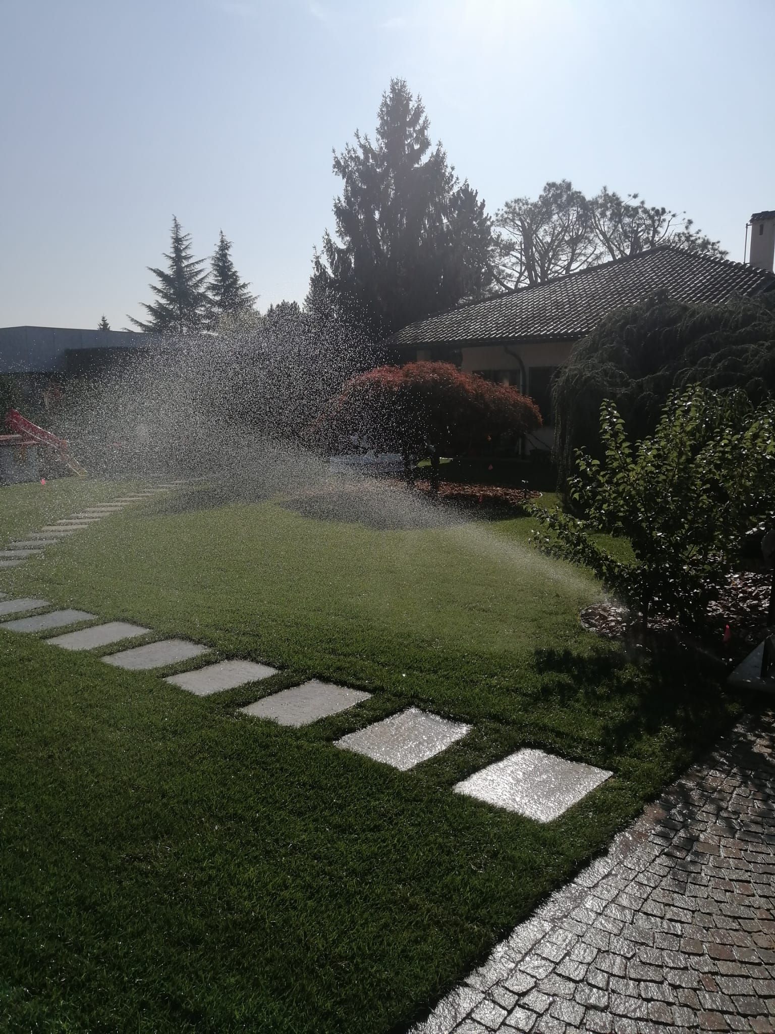 Un irrigatore da giardino spruzza acqua su un prato verde con pietre da calpestio, sotto un sole splendente.