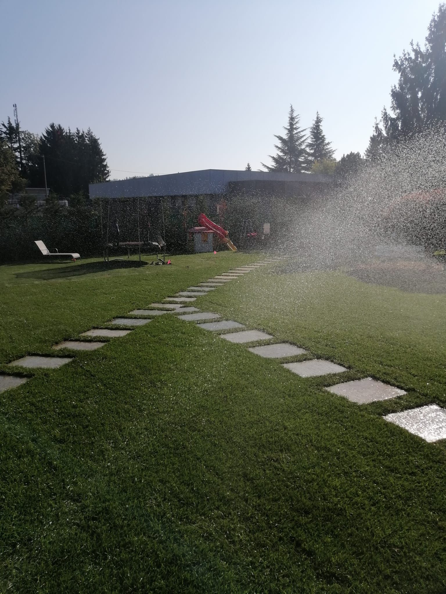 Prato nel cortile sul retro con pietre da calpestio e irrigatori che spruzzano acqua alla luce del sole