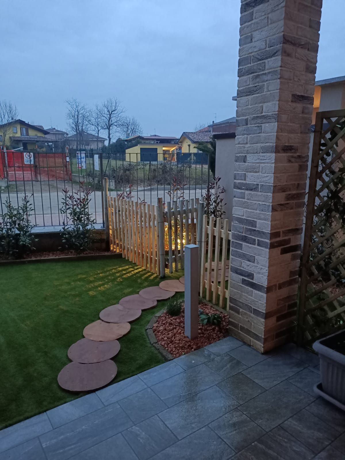 Patio sul retro con pavimentazione in pietra, prato, recinzione in legno e vista sul quartiere al crepuscolo.