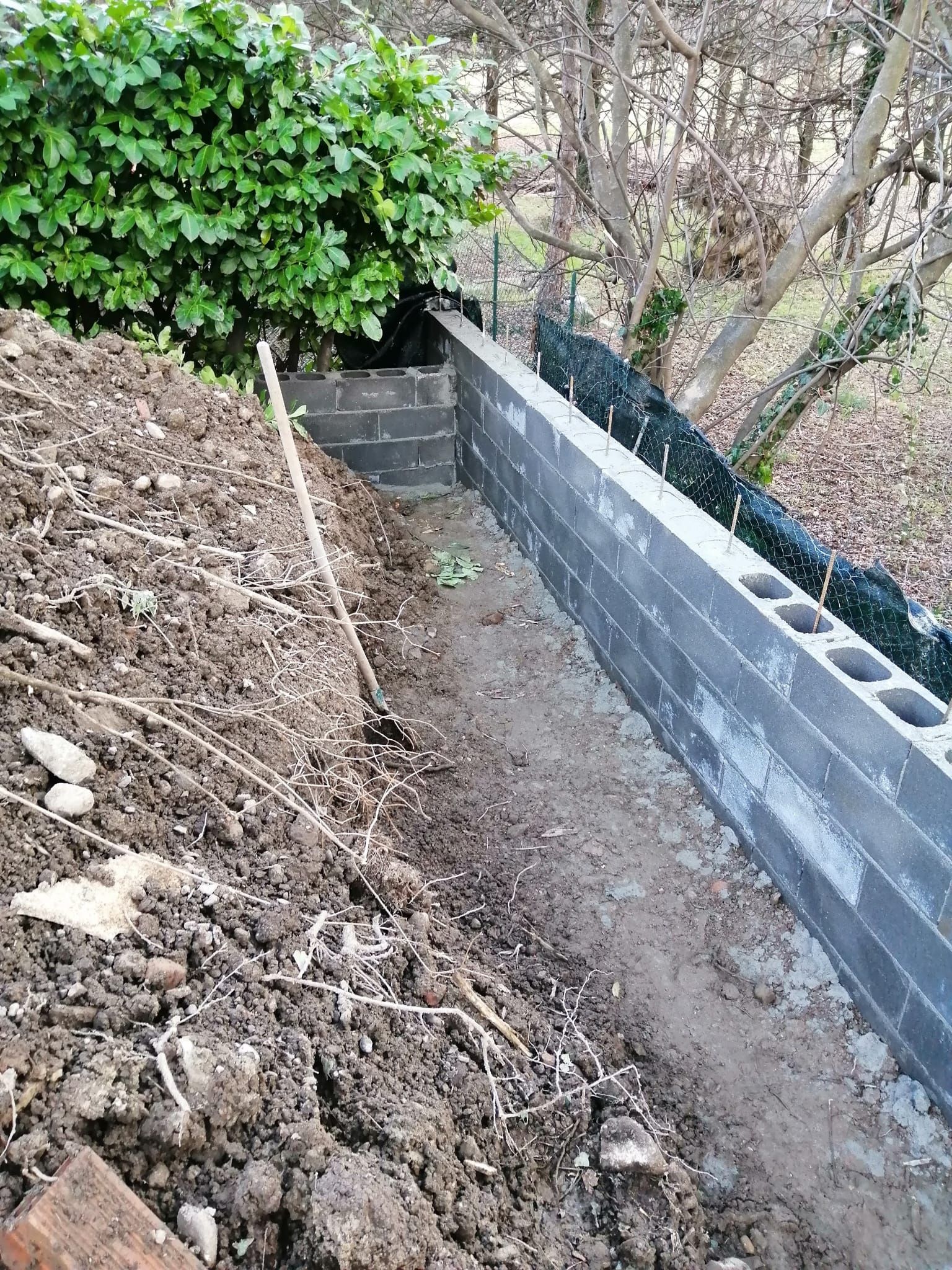 Stretto sentiero sterrato accanto a un muro di contenimento in blocchi di cemento grigi, con pendio di terreno e cespugli verdi.