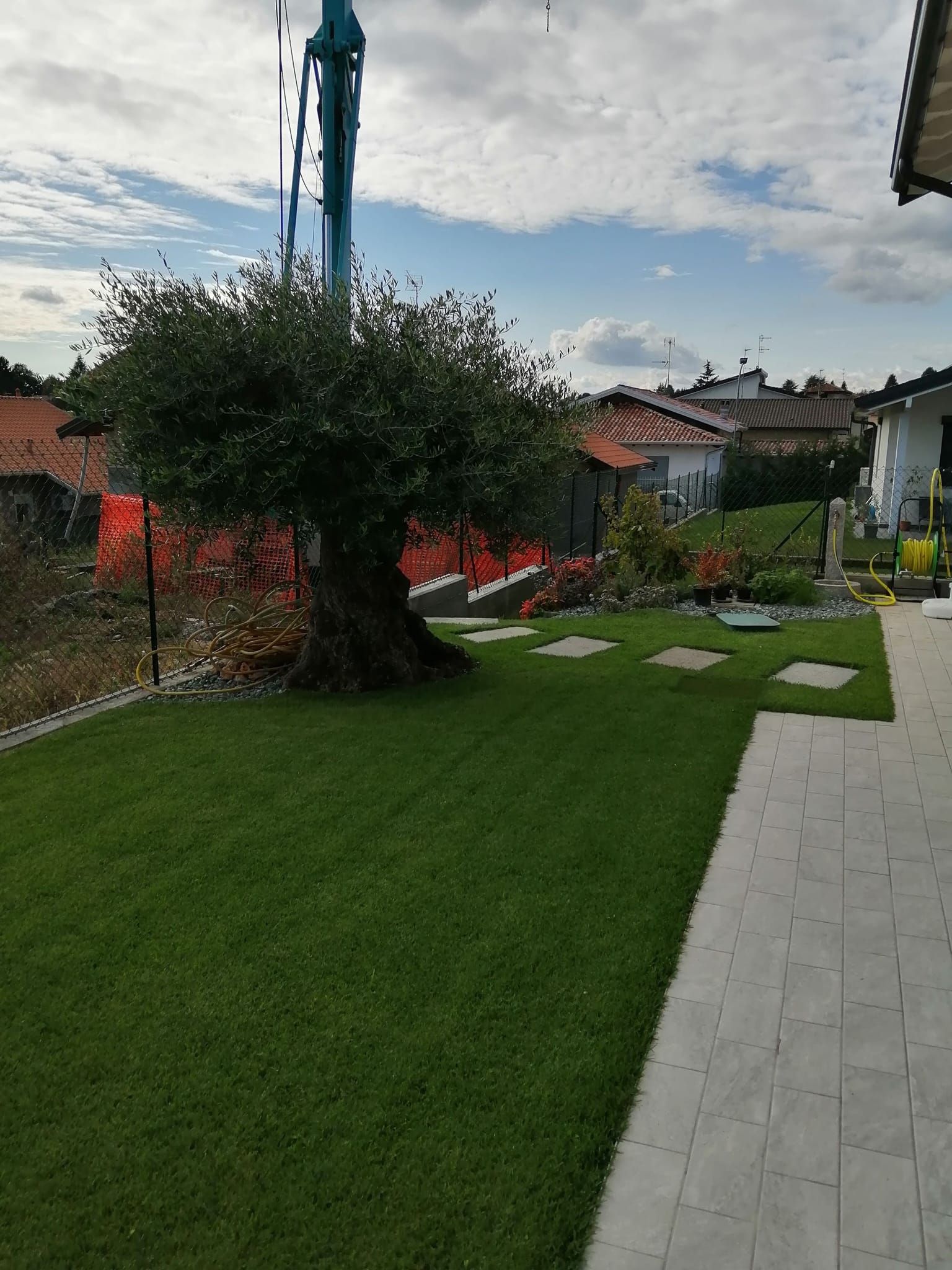 Giardino curato con prato verde, albero, sentiero in pietra e casa in costruzione sullo sfondo.