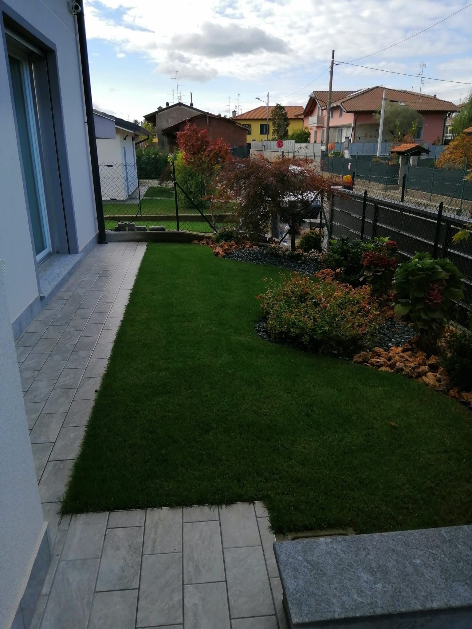 Cortile stretto sul retro con prato verde, aiuole fiorite e un vialetto pavimentato accanto alla casa e alla recinzione.