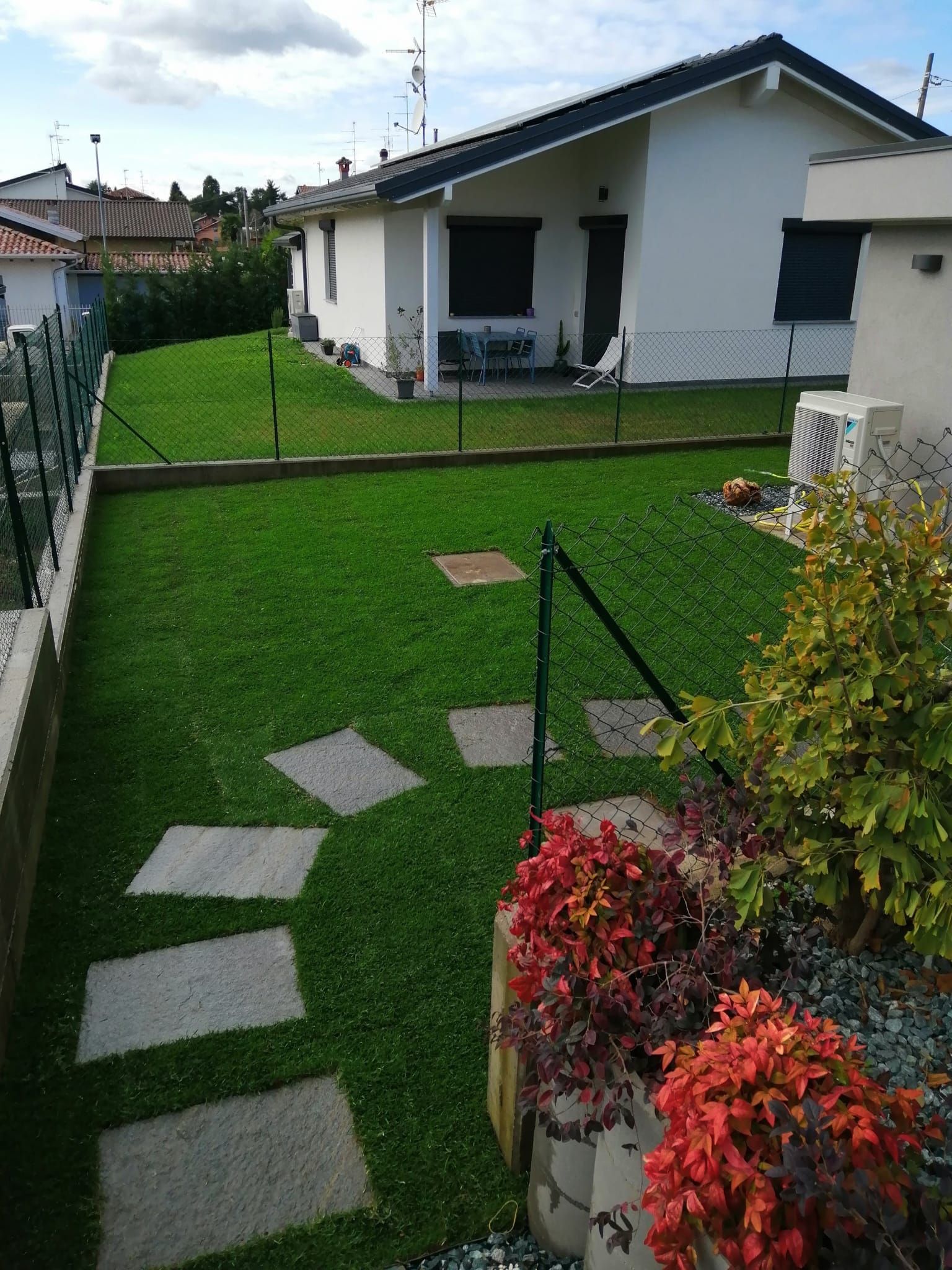 Giardino sul retro con prato verde, lastre di pietra, fiori in vaso e, sullo sfondo, il patio di una casa bianca.