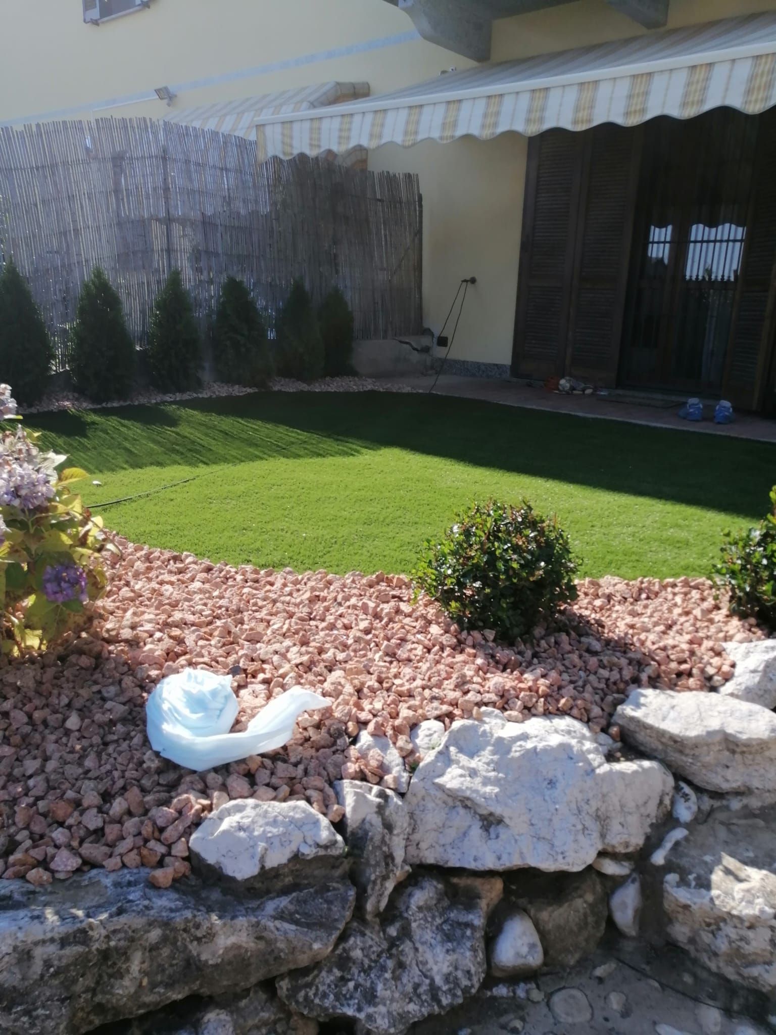 Giardino anteriore soleggiato con prato verde, bordo in pietra, arbusti e, sullo sfondo, il portico di una casa.