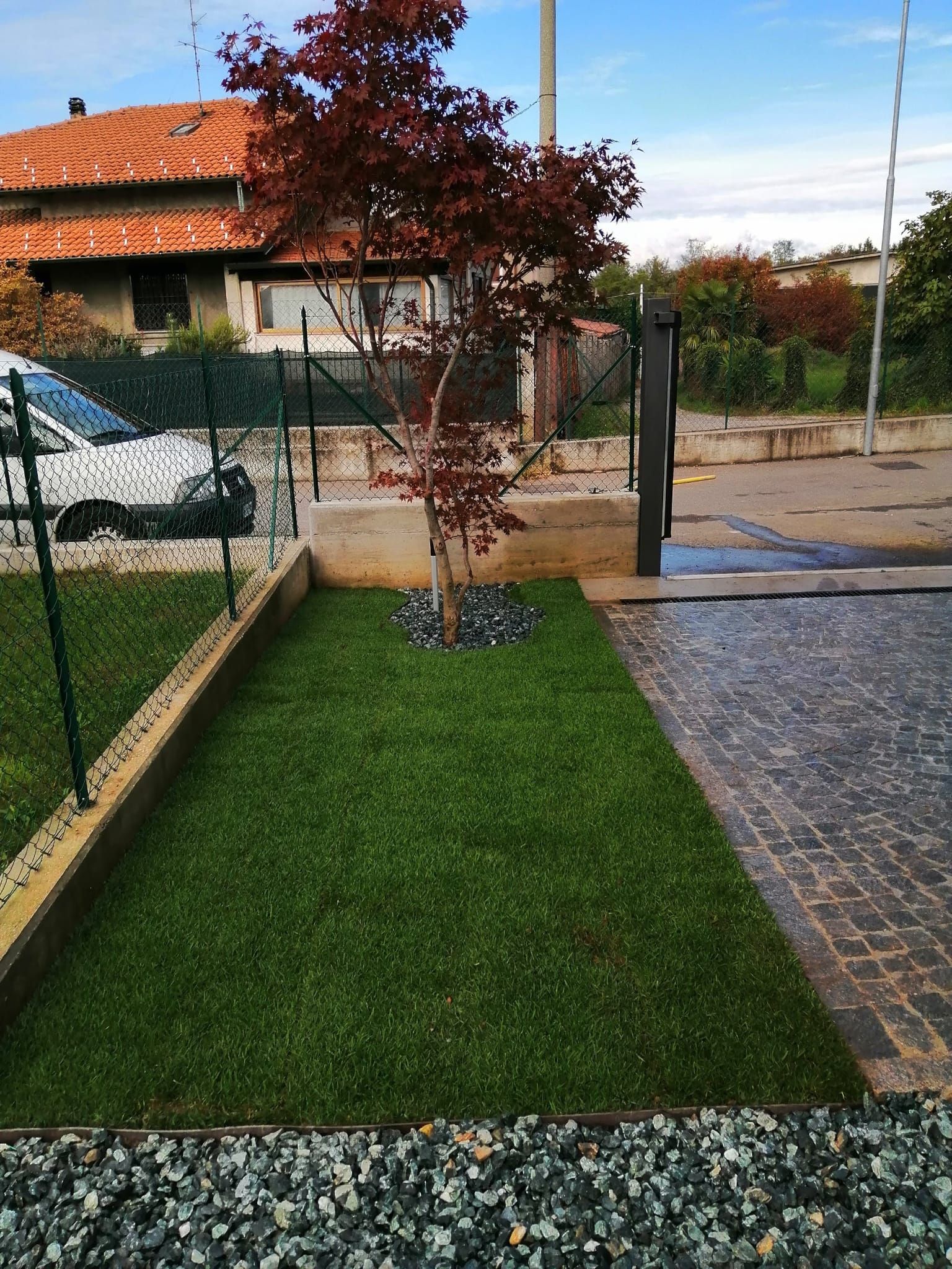 Cortile anteriore con prato verde, piccolo albero dalle foglie rosse, sentiero in pietra e, sullo sfondo, casa con tetto di tegole.