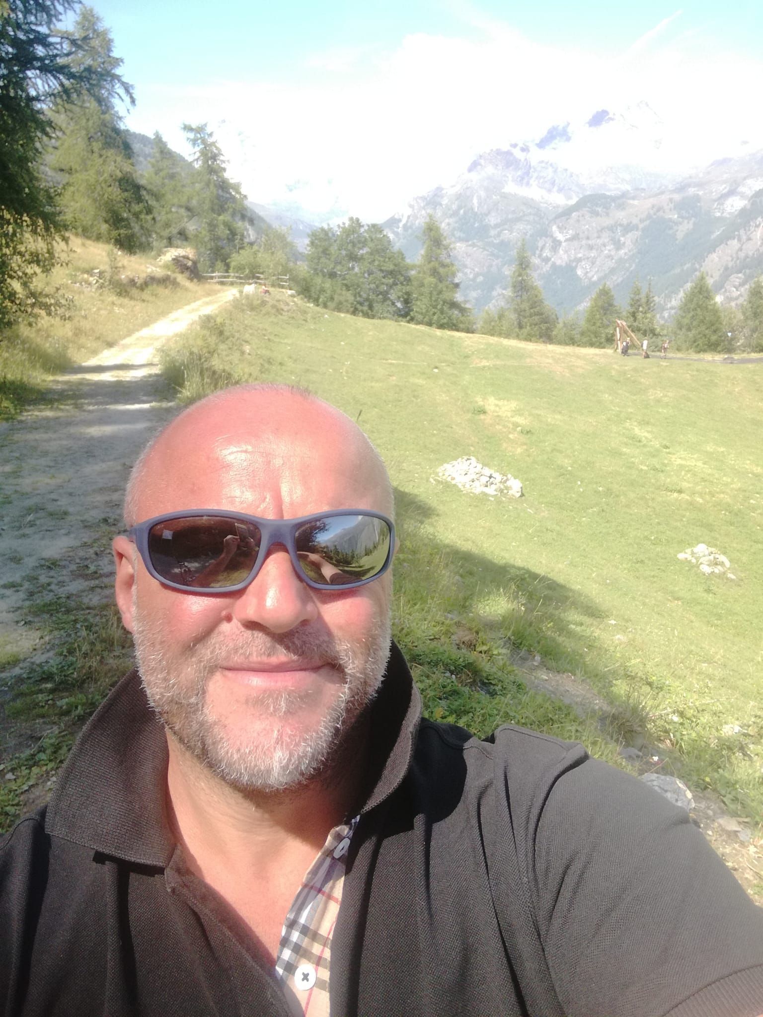 Un uomo si scatta un selfie su un sentiero di montagna, indossando occhiali da sole, con cime innevate e alberi sullo sfondo.
