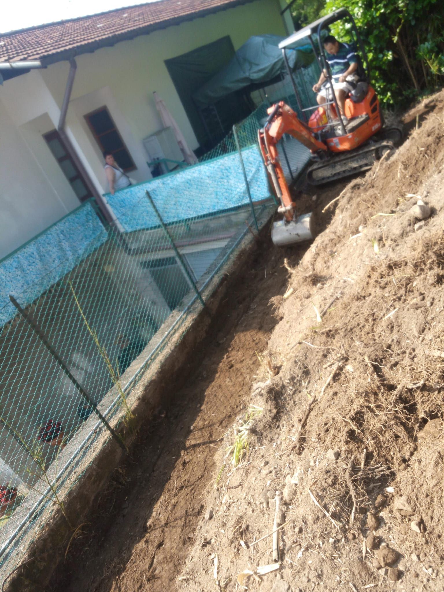 Un escavatore arancione sta scavando una trincea accanto a una casa con una recinzione blu.