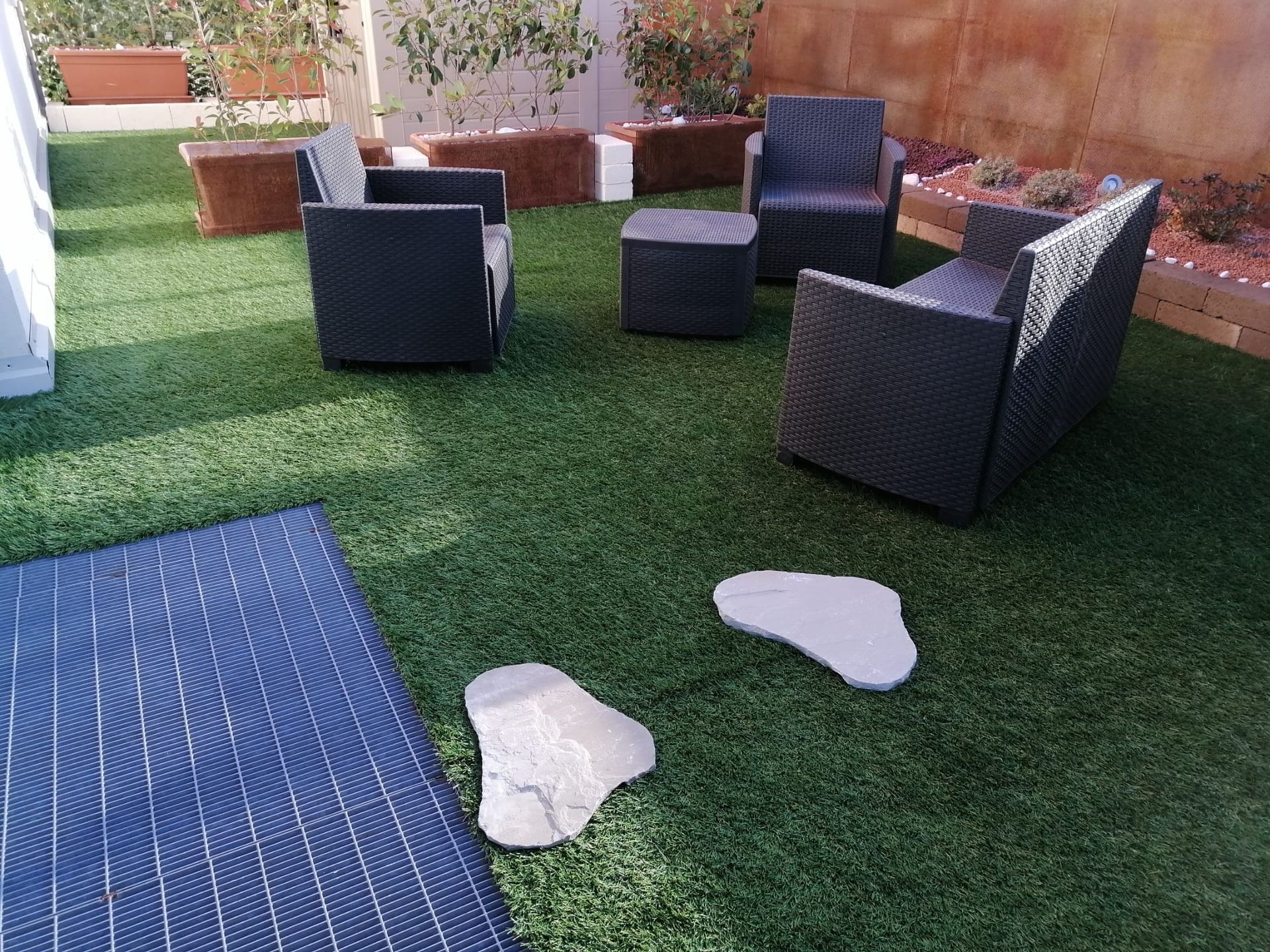 Patio esterno con sedie in vimini nere su erba artificiale, un tappetino blu e due pietre bianche da calpestio.