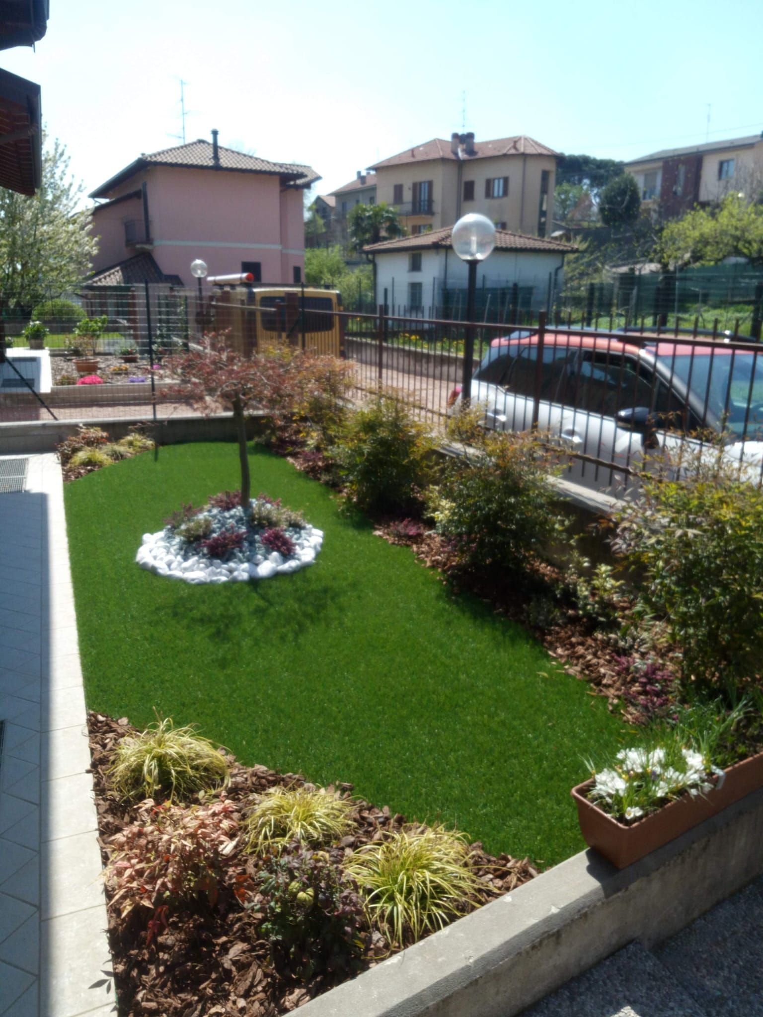 Piccolo giardino antistante la casa con prato verde, aiuole e un'auto parcheggiata lungo una strada residenziale.