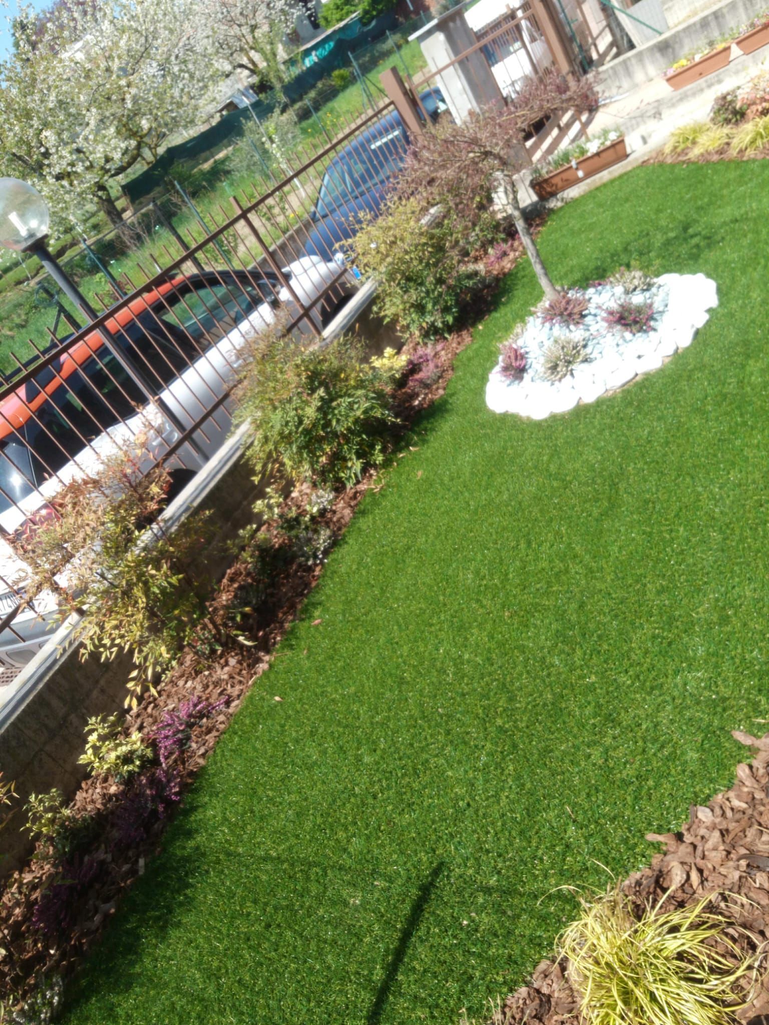 Vista inclinata di un cortile paesaggistico con prato verde, arbusti, aiuola di fiori bianchi e un vialetto recintato vicino a un edificio.