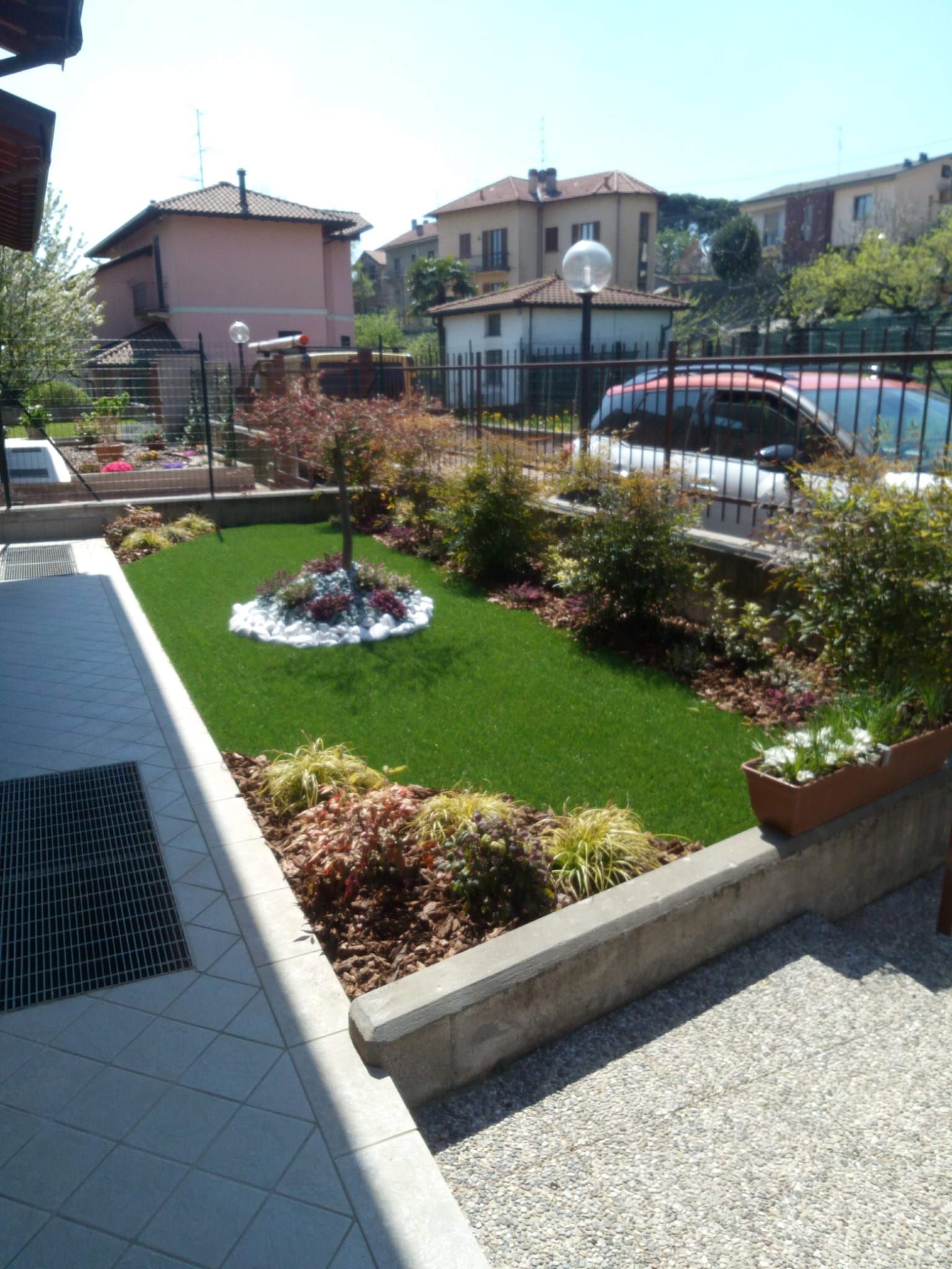 Piccolo giardino anteriore ben curato con prato, arbusti e un vialetto accanto ad auto e case parcheggiate.