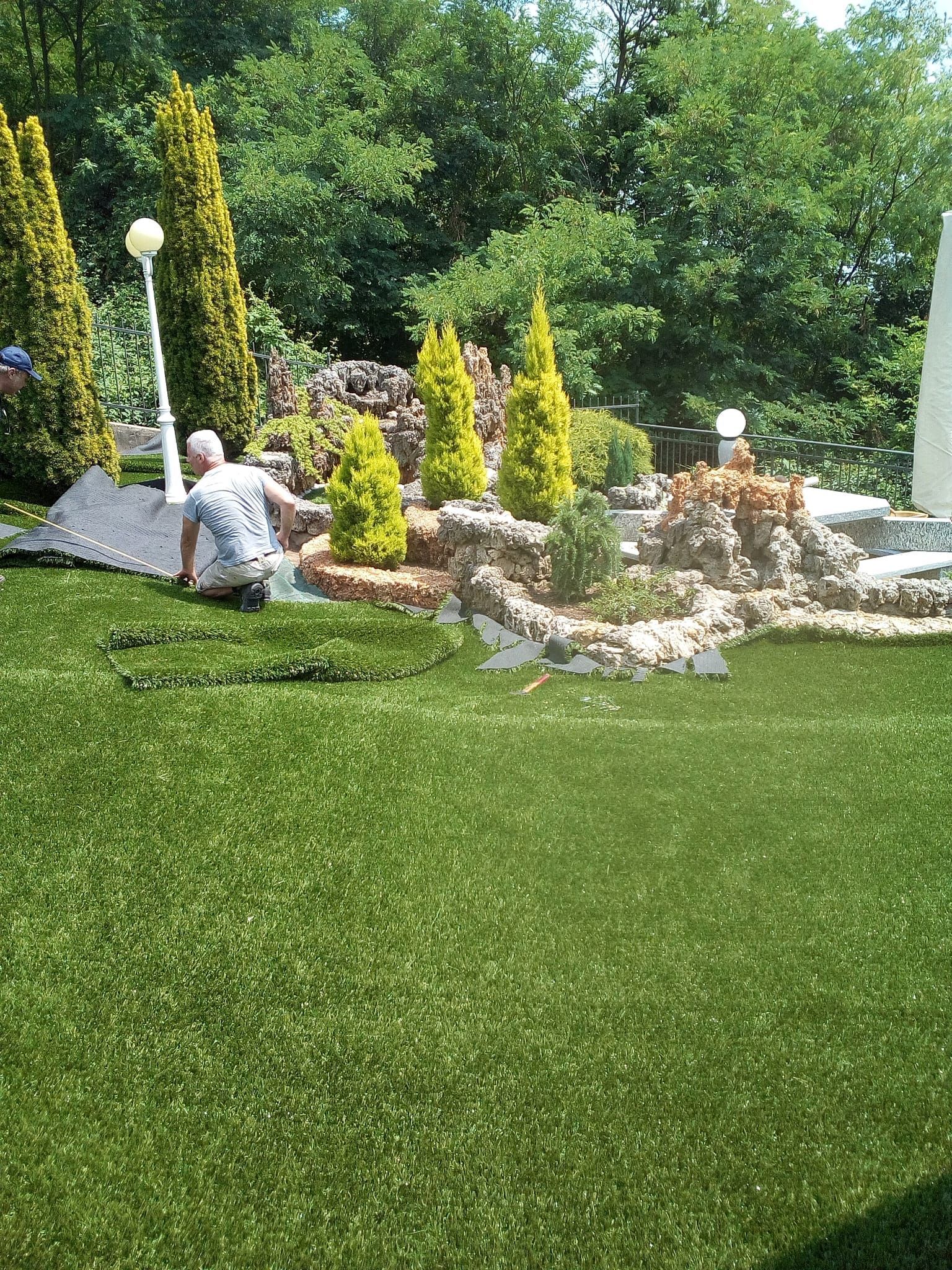 Una persona con un cappello bianco è seduta in un giardino curato con rocce, arbusti e prato verde.