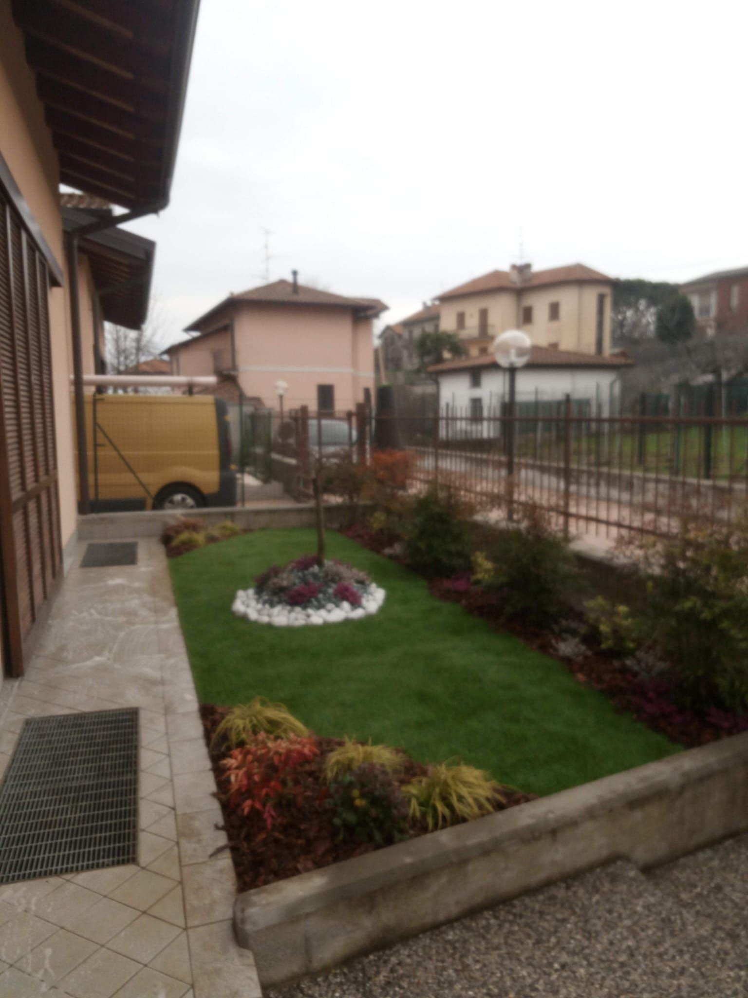 Piccolo giardino anteriore ben curato con prato verde, aiuola e sentiero in pietra, adiacente alla casa e alla strada recintata.
