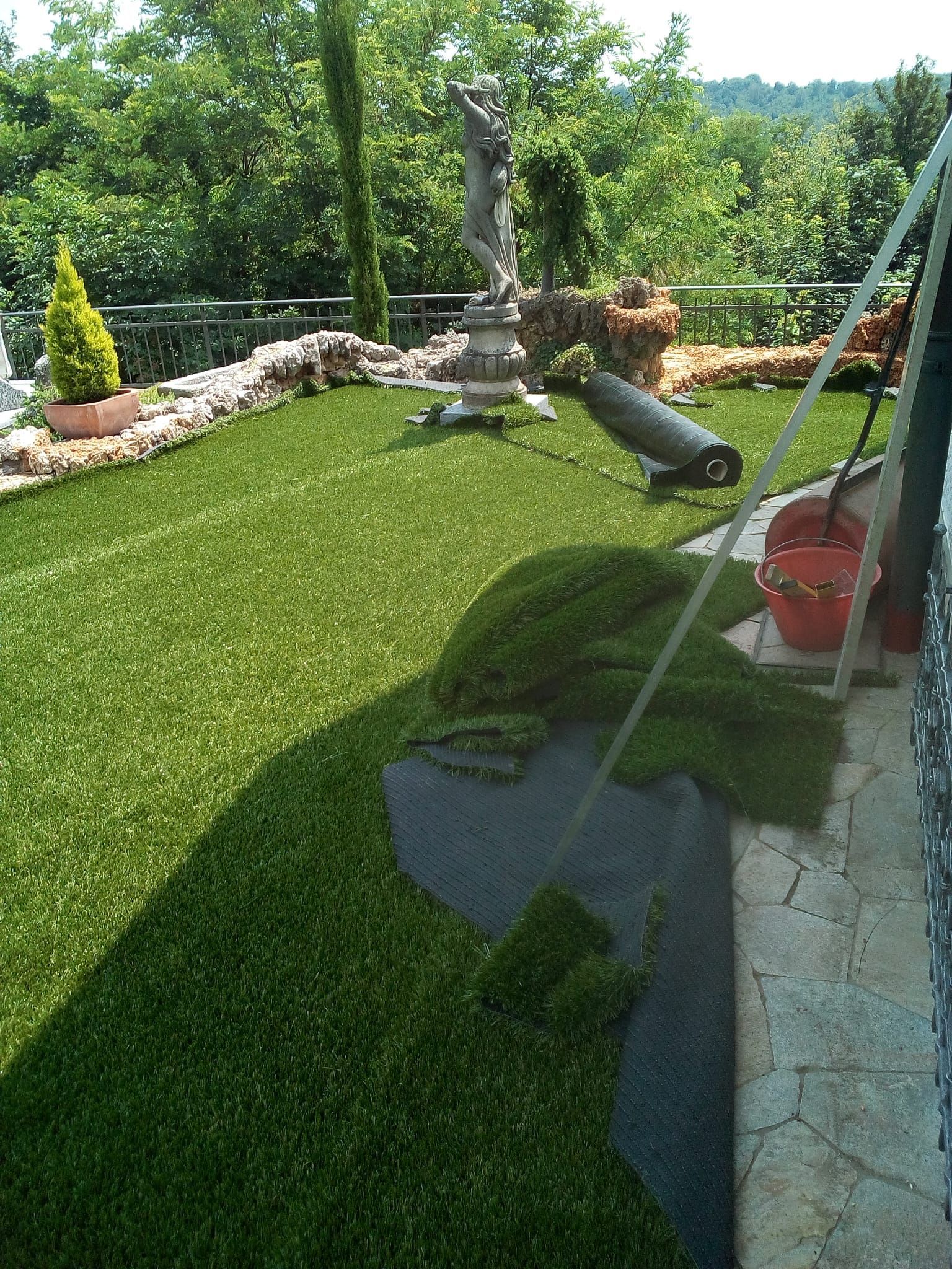 Cortile soleggiato con prato verde lussureggiante, bordo in pietra, statua e tubo da giardino che spruzza acqua dal lato destro.