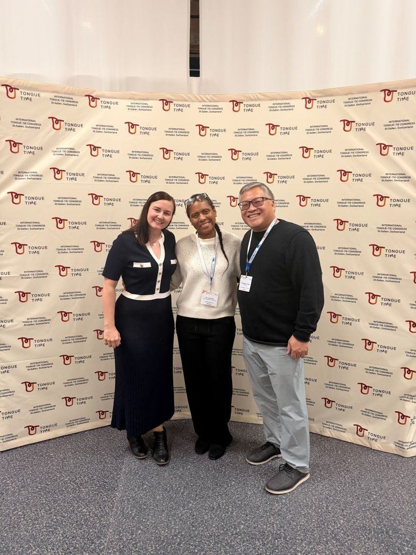 Three people smiling at a branded event backdrop, standing together on carpeted flooring.