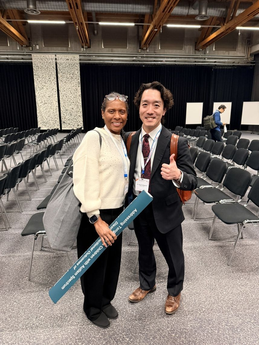 Two people smiling and posing in an empty auditorium, holding a teal sign and a medal.