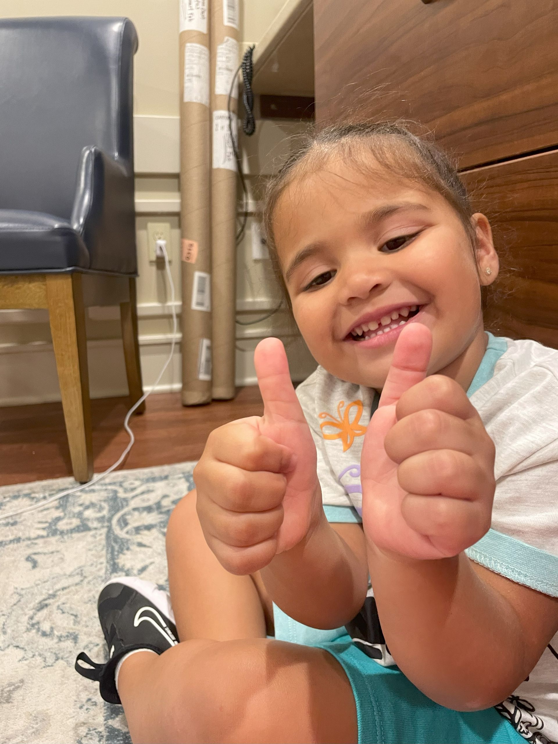 Girl smiling, giving double thumbs-up; seated on floor. Wearing teal shorts, black shoes, and a white t-shirt.