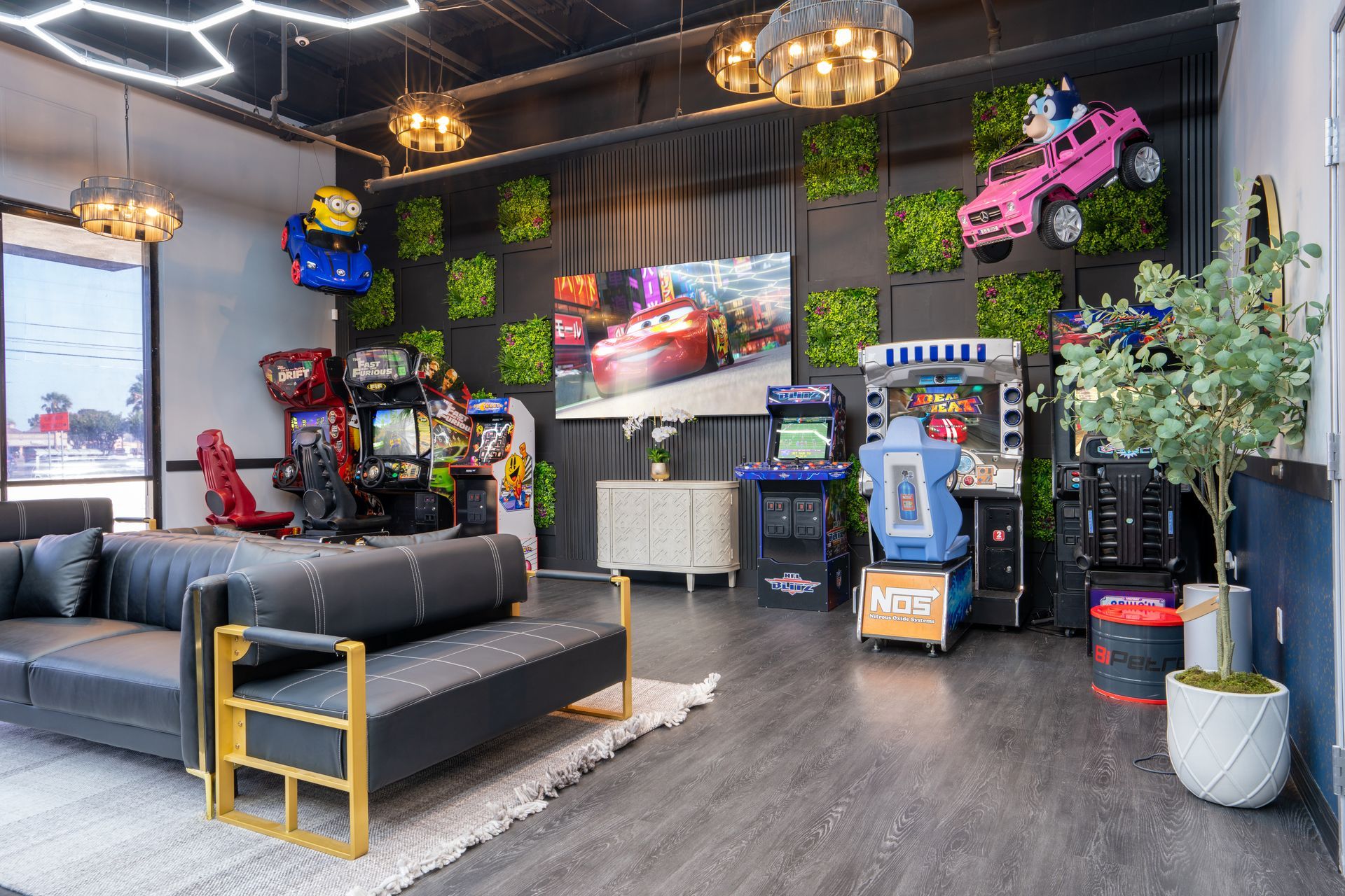 Modern office lounge with black sofa, TV, arcade machines, hanging lights, plants, and a pink toy car on the wall