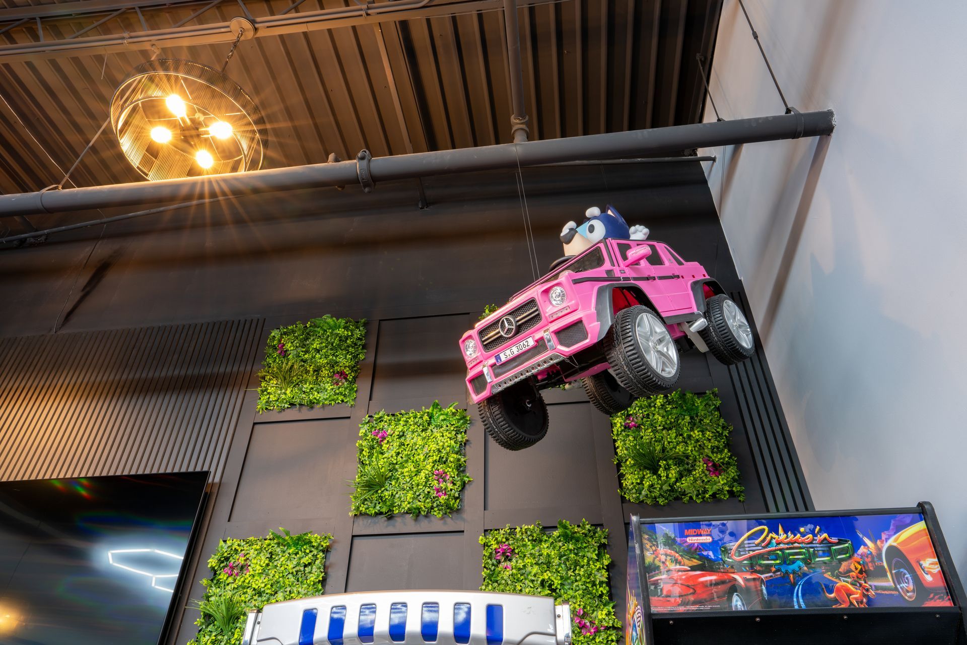 Pink toy car mounted on a wall in an industrial-style interior with green plant panels and a hanging light