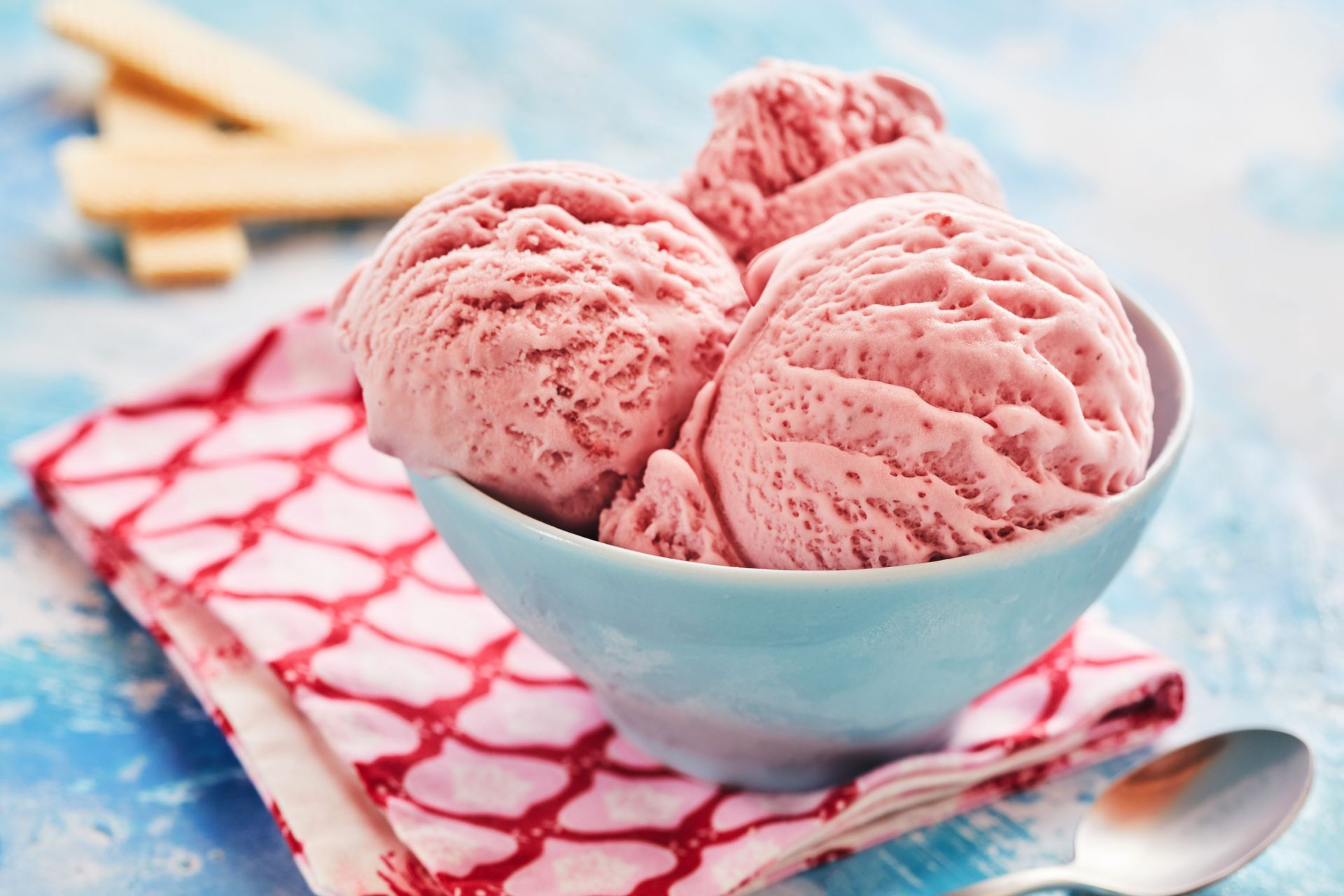 Three scoops of pink strawberry ice cream in a blue bowl on a patterned napkin, with spoons nearby