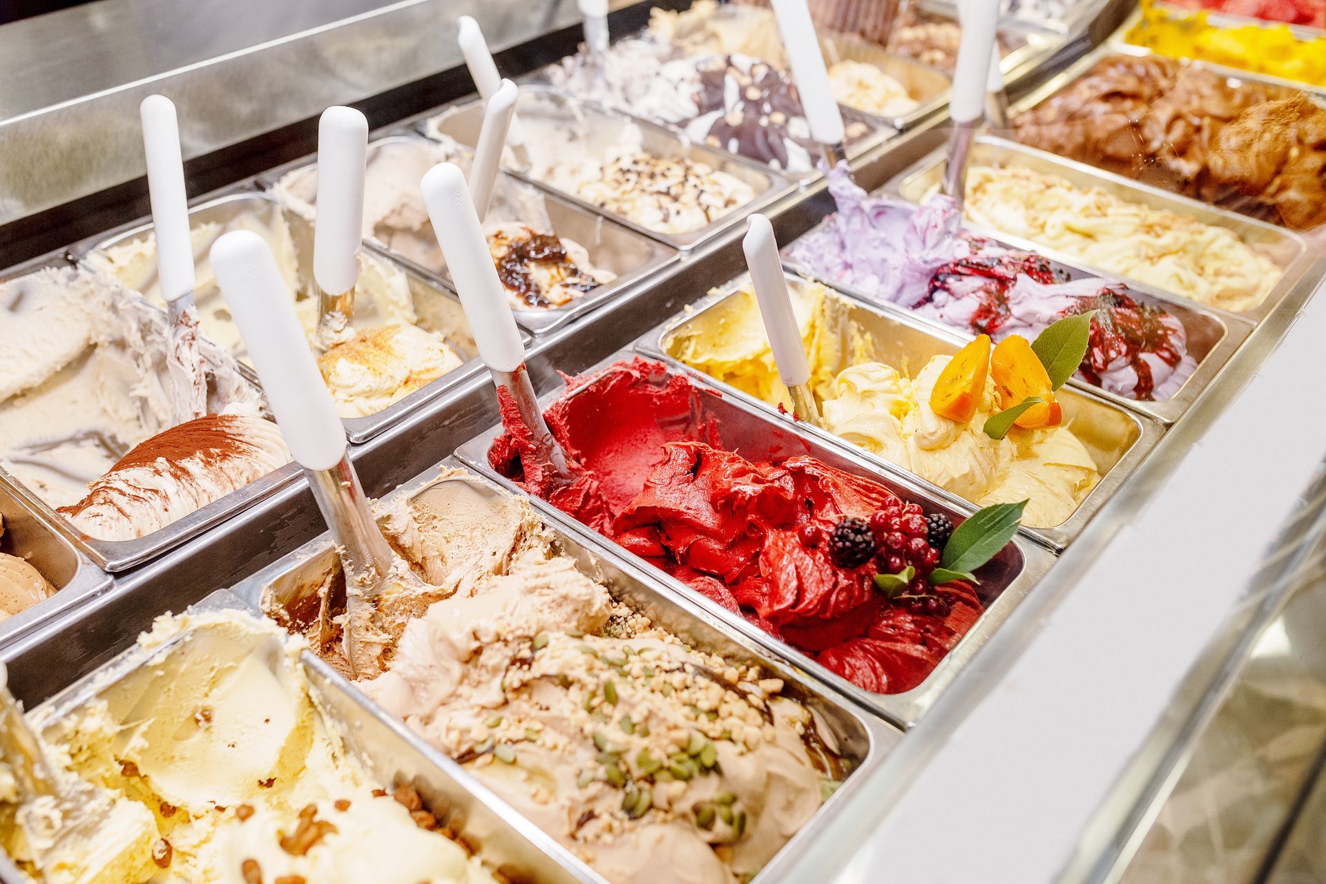 Une vitrine remplie de rangées de glaces colorées dans des récipients métalliques, chacun accompagné d'une cuillère de service.