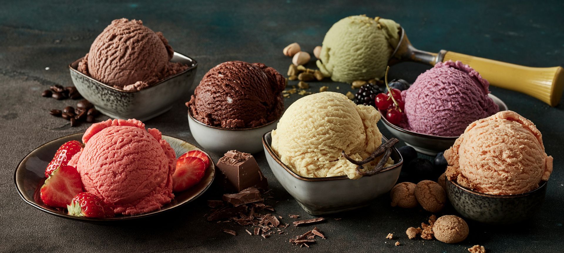 Six boules de glace colorée dans des petits bols, disposées sur une table avec des fraises, des baies, du chocolat et des noix.