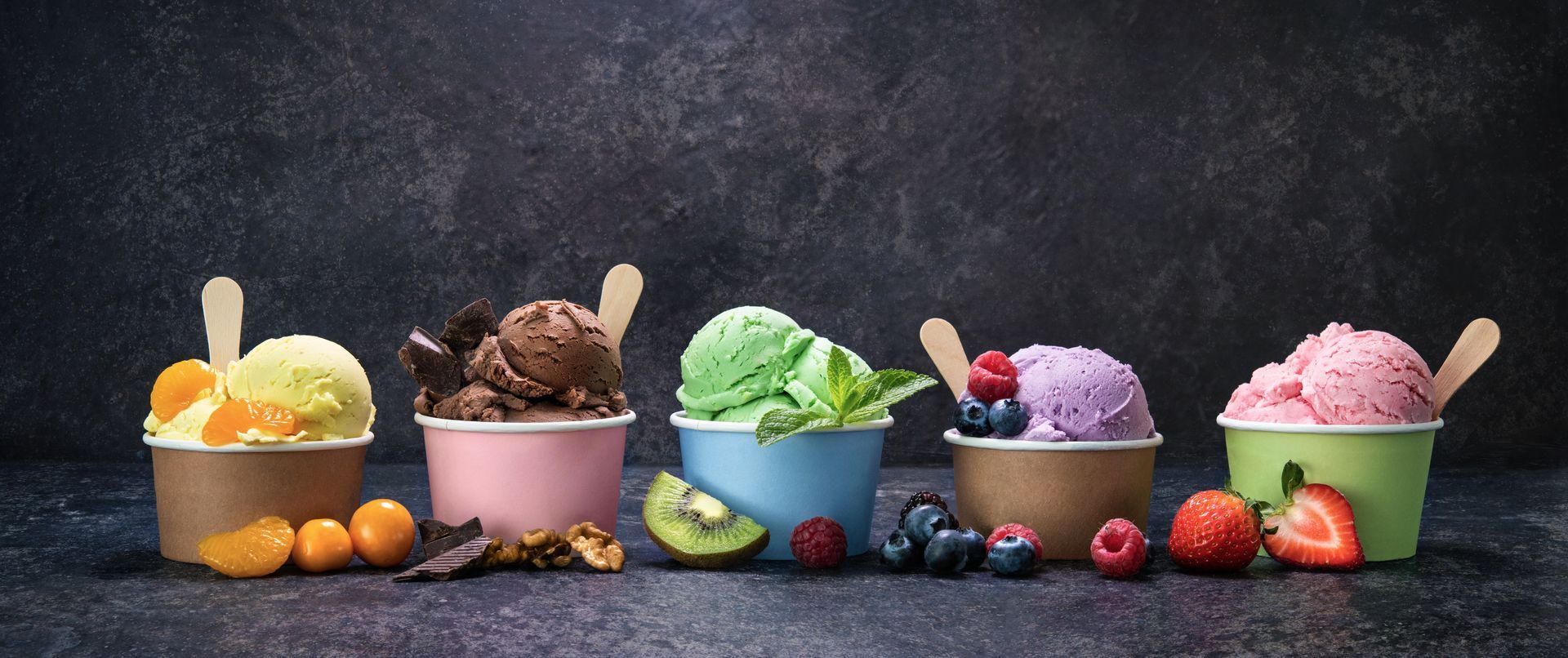 Cinq gobelets en papier colorés remplis de glaces aux parfums différents, chacun accompagné d'une cuillère en bois posée sur une surface texturée foncée.