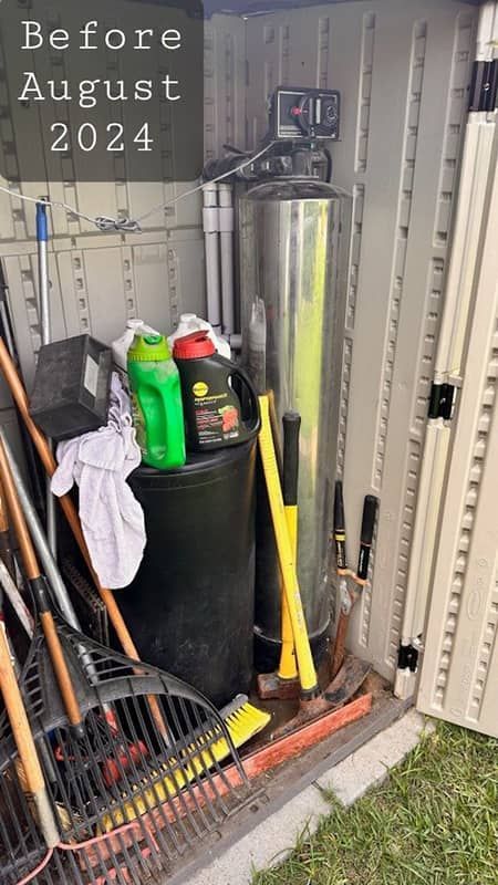 Storage closet with tools, gas cans, and a black barrel beside a water heater, labeled “Before August 2024”