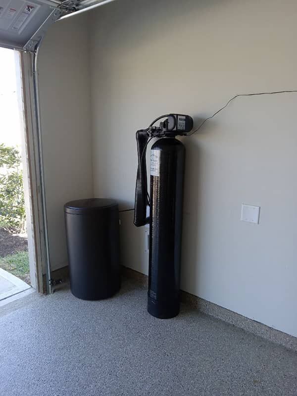 Indoor garage corner with a black water softener tank and cylindrical storage bin beside a wall.