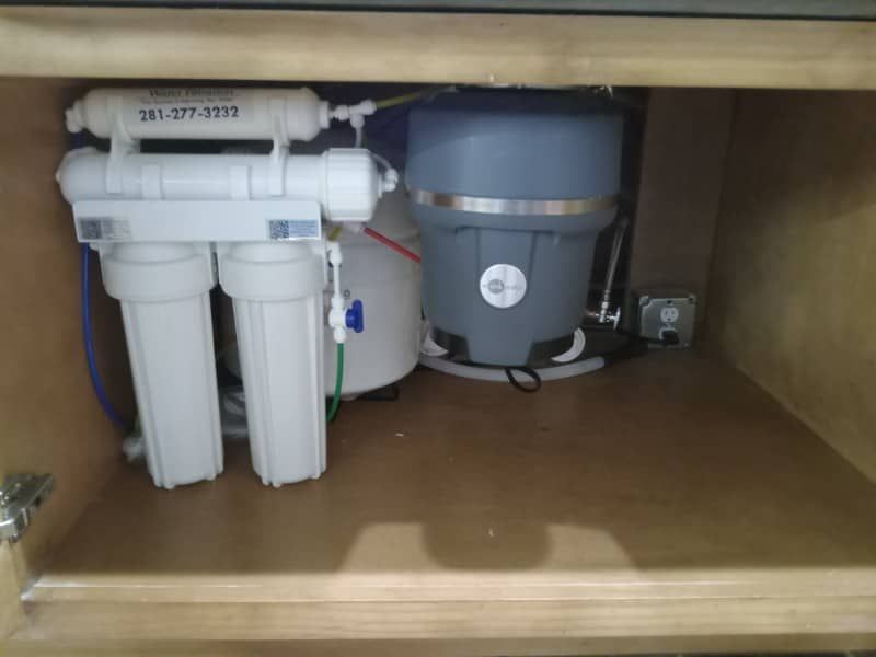 Under-sink water filtration system with white filters and a gray tank in a wooden cabinet