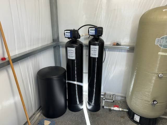 Water softener tanks and a black resin tank in a utility room beside a beige pressure tank.