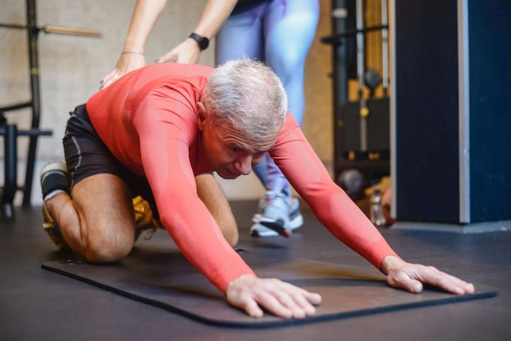 Man in red shirt stretching on mat with trainer's hands on back in gym.