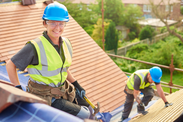 Two roof workers in blue hard hats and safety vests installing shingles on a rooftop