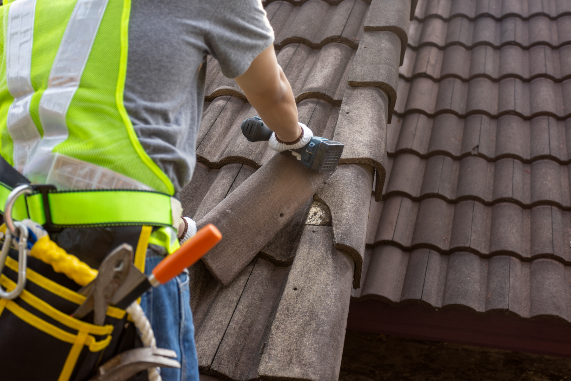 Worker repairing a roof, cutting brown roof tiles with a hand saw while wearing a safety harness.