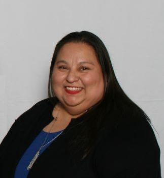 Diasy smiling, wearing a blue shirt and black sweater against a white backdrop.