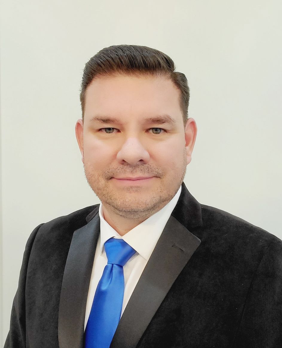 Edgar J Hernandez in black suit and blue tie, smiling, against a white background.