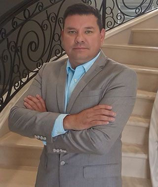 Man in gray suit with arms crossed, standing on a staircase, blue shirt visible, neutral expression.