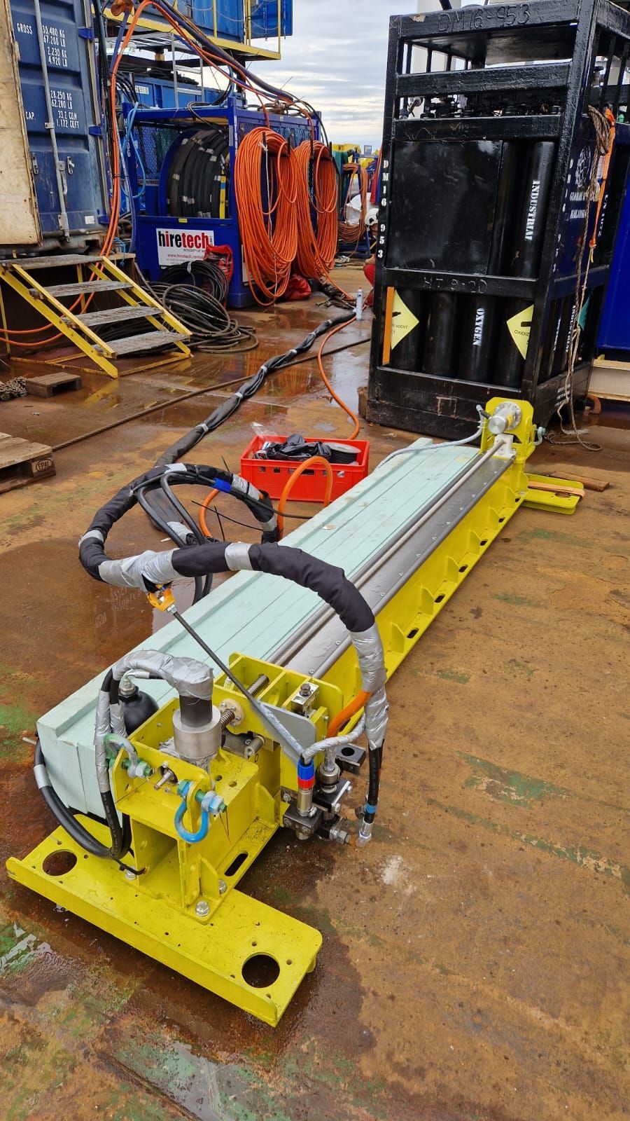 Yellow industrial machine with black hoses on a ship deck near other equipment.