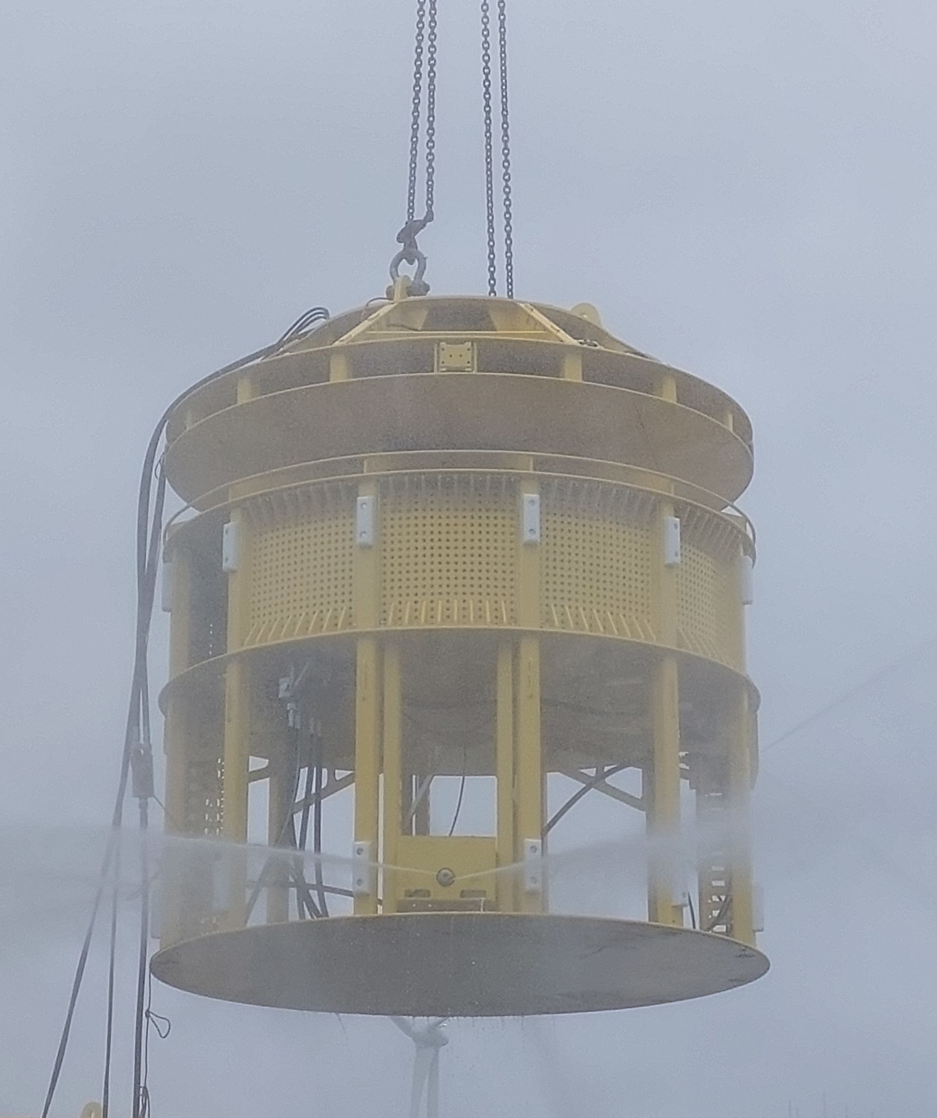Yellow cylindrical object, possibly machinery, suspended by a crane in misty conditions.