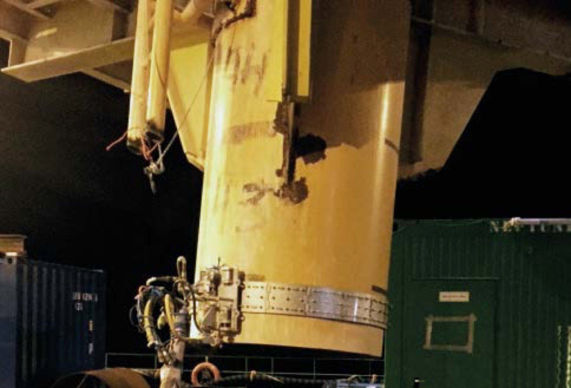 Damaged yellow crane component against dark background. Metal panels, bolts, and damage visible.