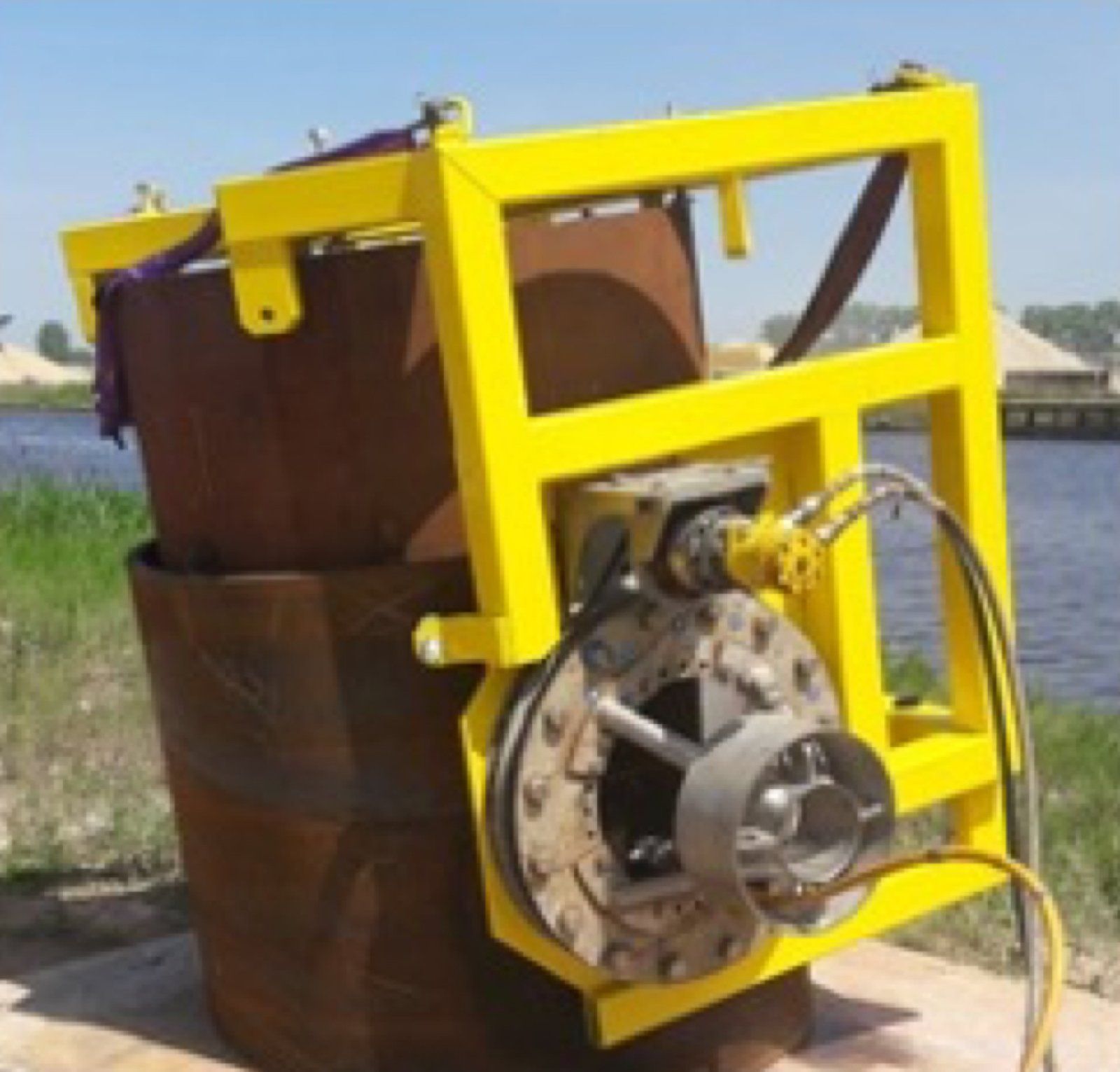 Yellow metal frame apparatus holding a rusty metal drum, outdoors near water.