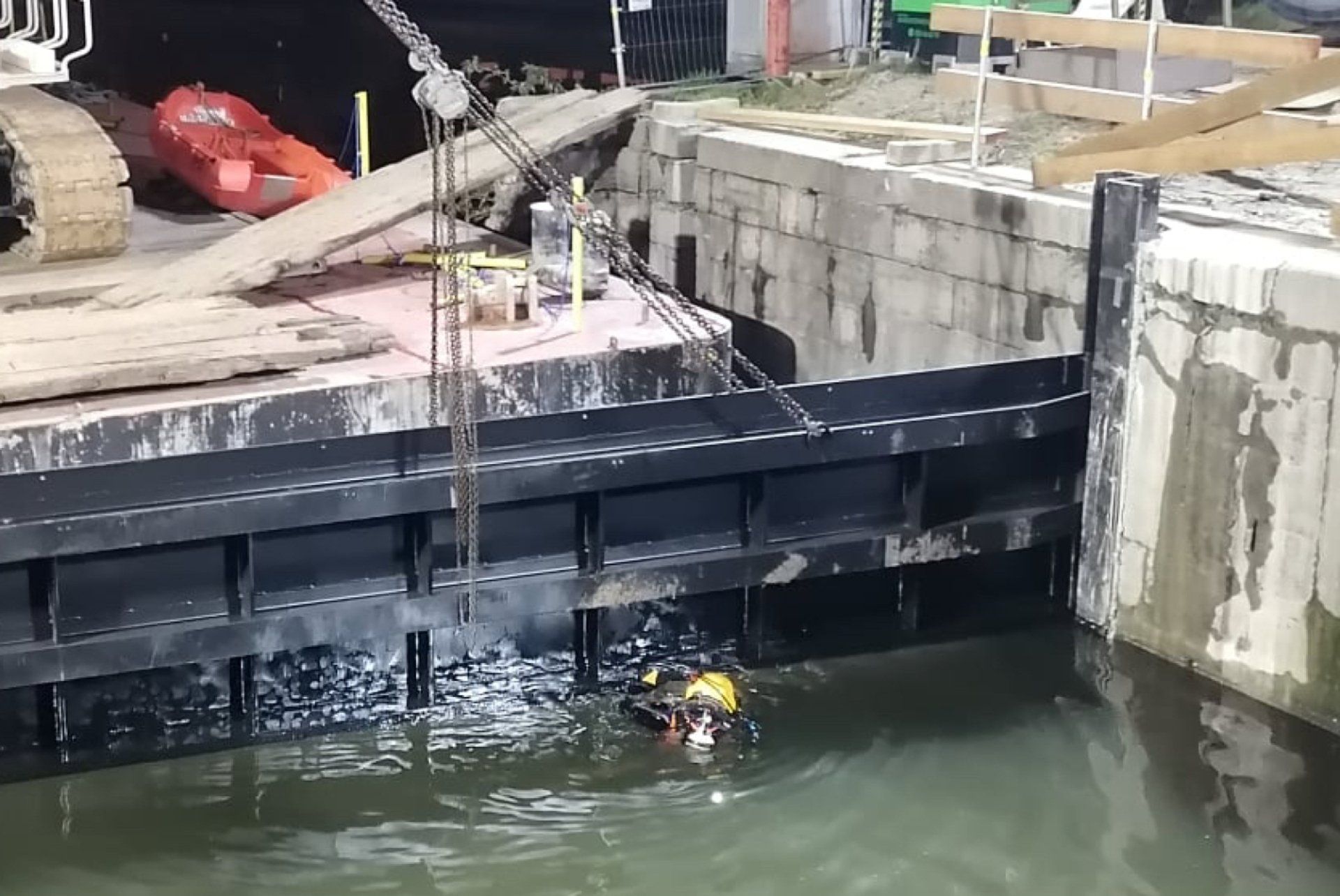 Diver inspecting a large black gate in a canal. Construction equipment visible. Water is murky.