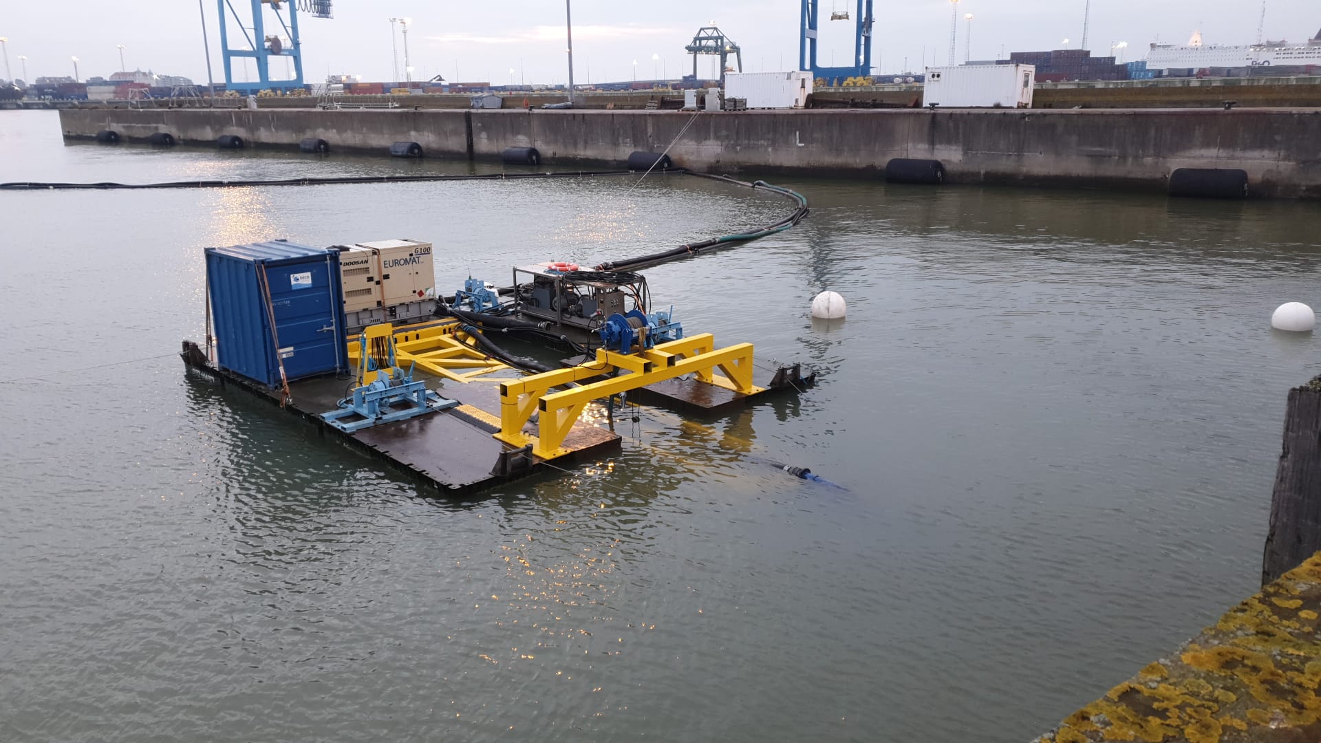 Oil skimmer barge in harbor, collecting oil. Blue, yellow, and black structures on water.