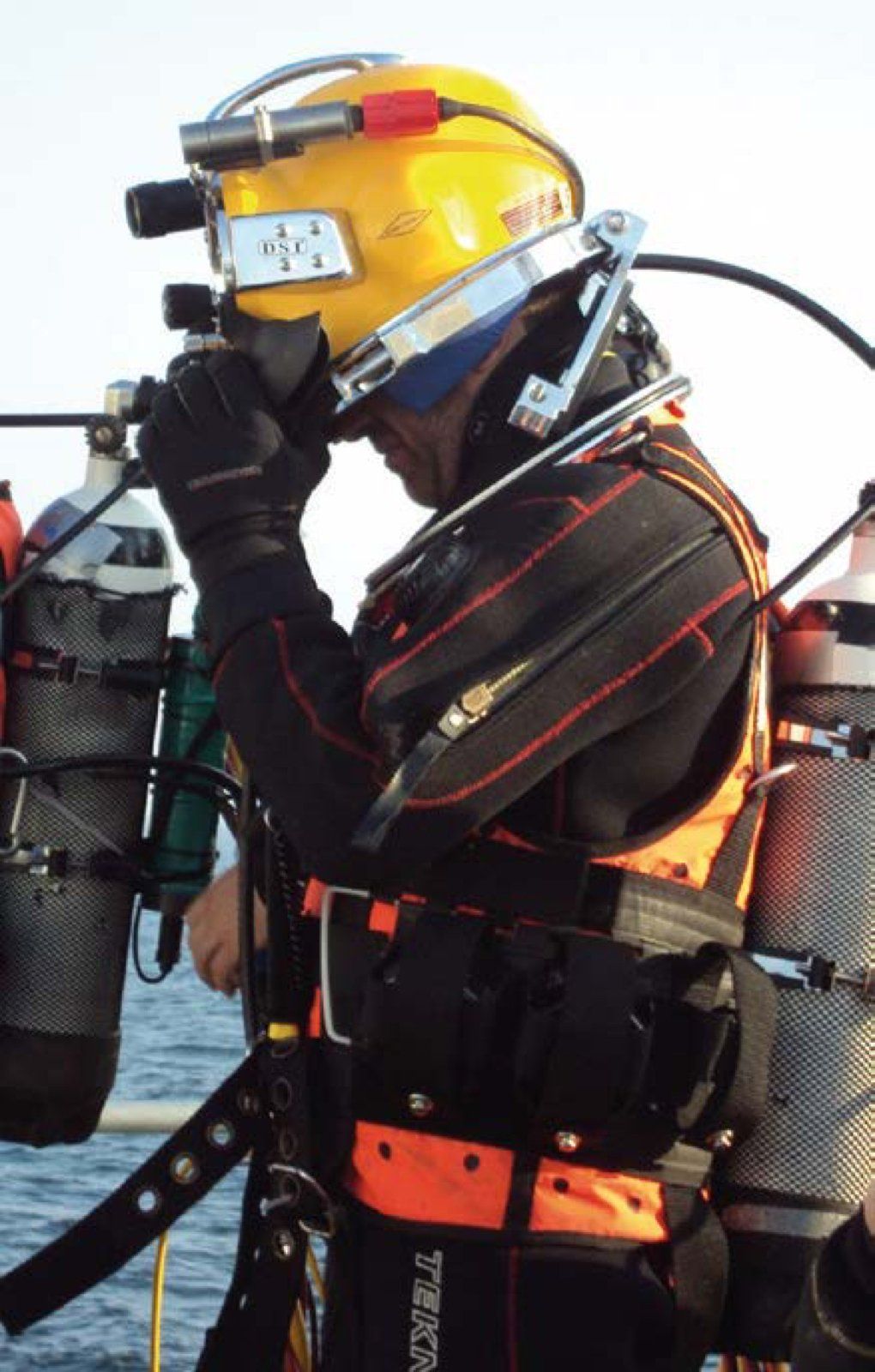 Diver in black and orange drysuit, yellow helmet, and diving equipment, on a boat deck, looking at something.