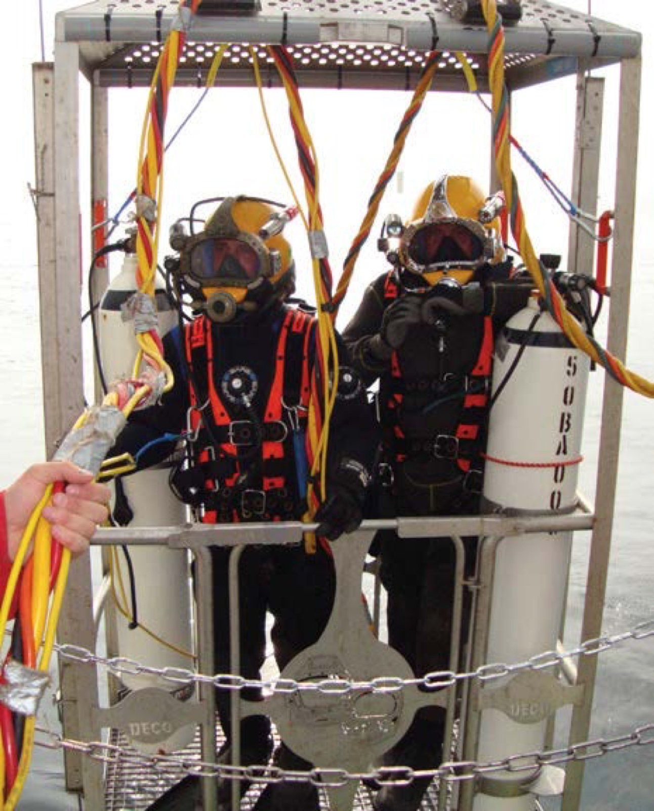 Two divers in diving suits in a metal cage, with air tanks, connected by cables.